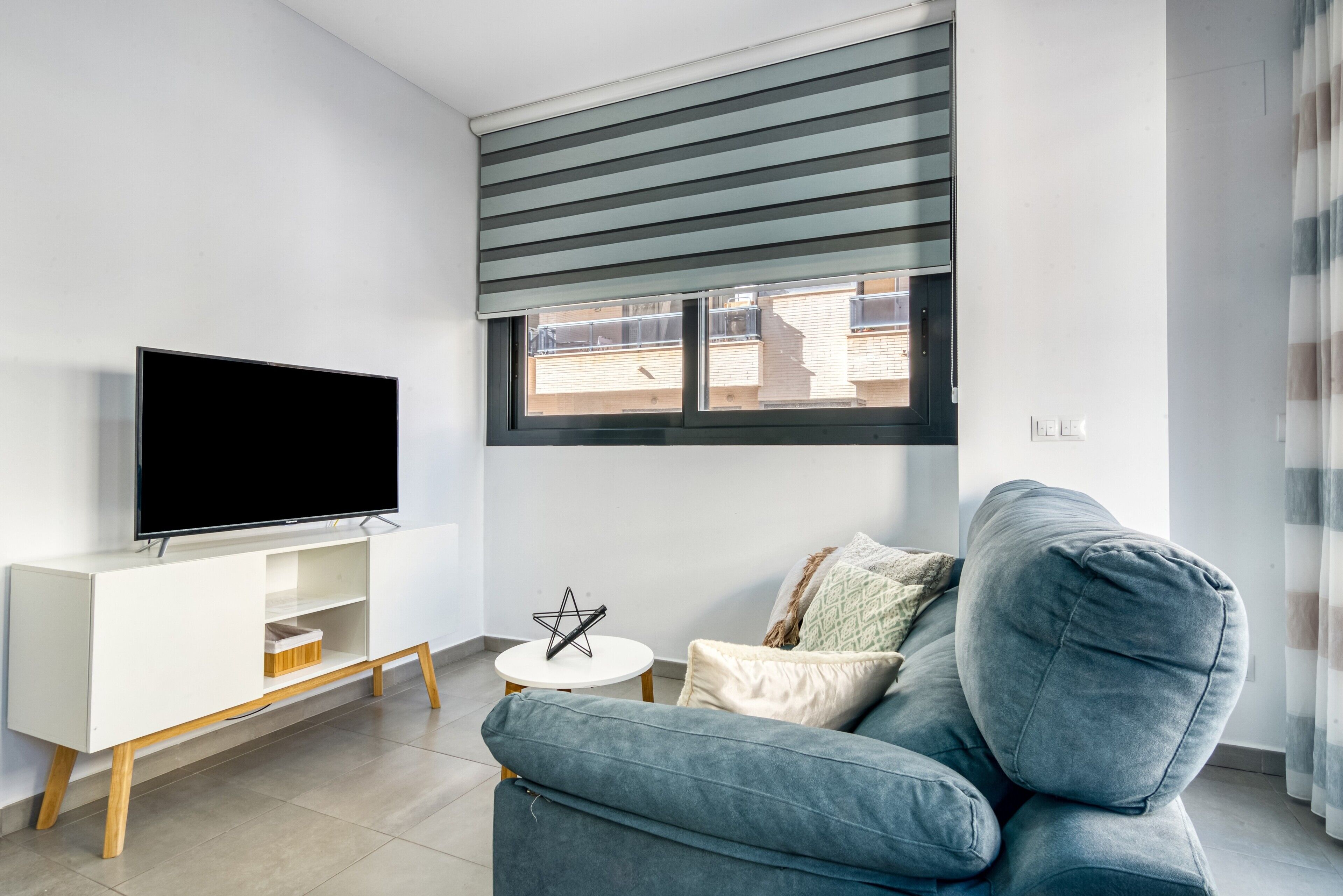 A modern living room with a flat-screen TV on a white TV stand, a grey sofa, and a small white table with a decorative item.
