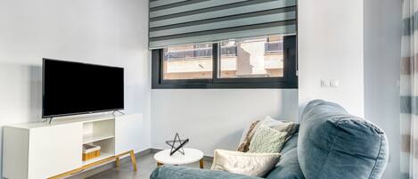 A modern living room with a flat-screen TV on a white TV stand, a grey sofa, and a small white table with a decorative item.