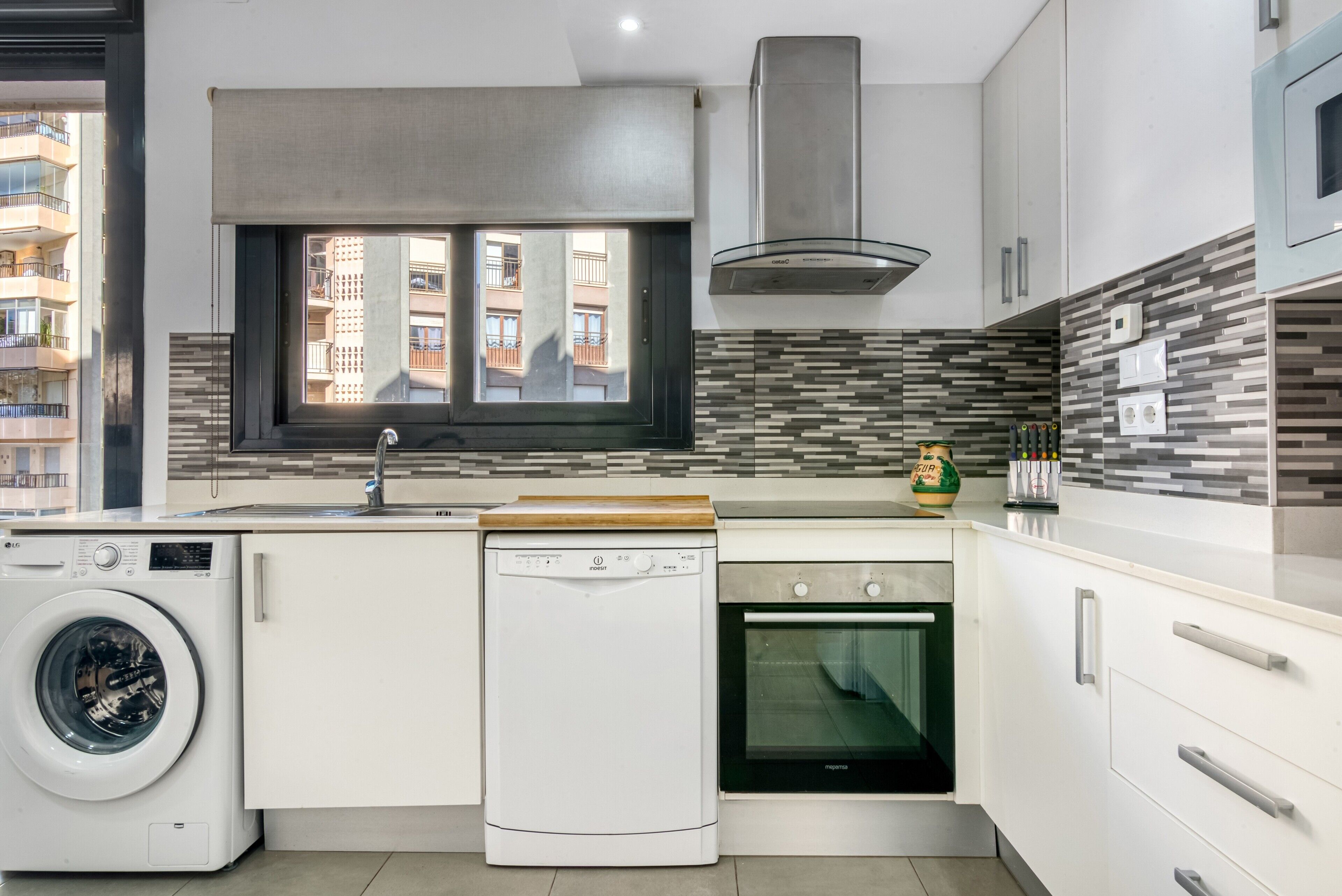 A compact kitchen with a washing machine, dishwasher, and oven, a window with a view of buildings, and a tiled backsplash.