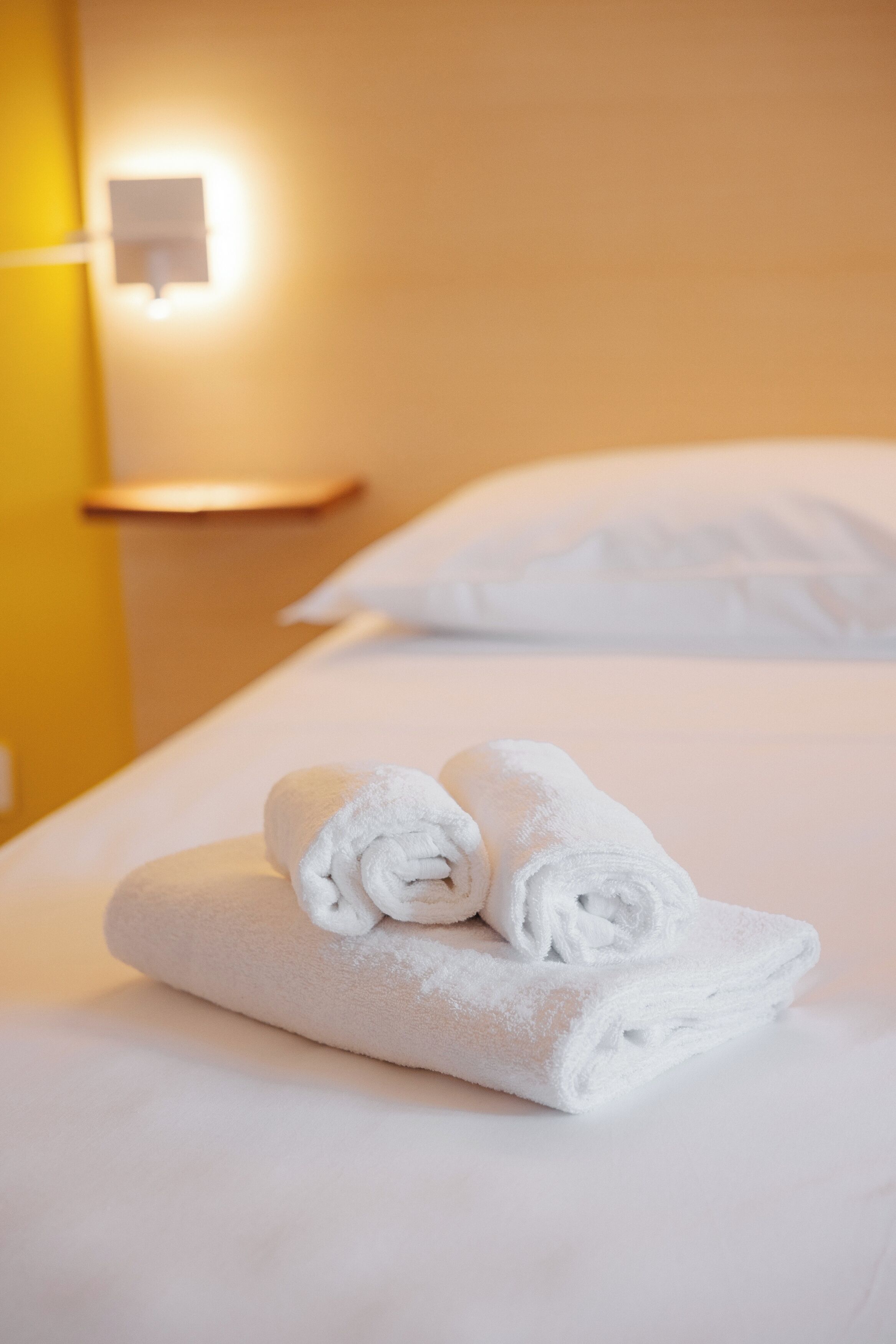 A neatly made bed with two rolled towels and a wall-mounted light fixture.