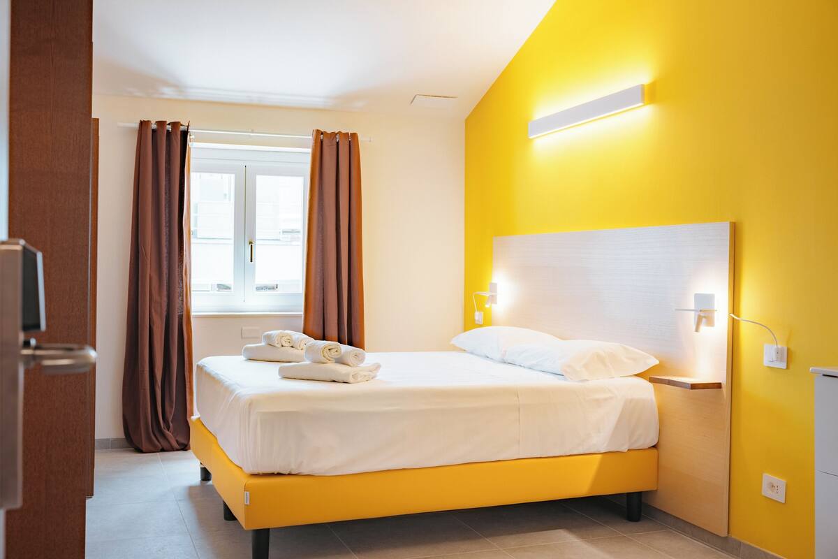 A hotel room with a bed, curtains, a window, and a wall-mounted light fixture.