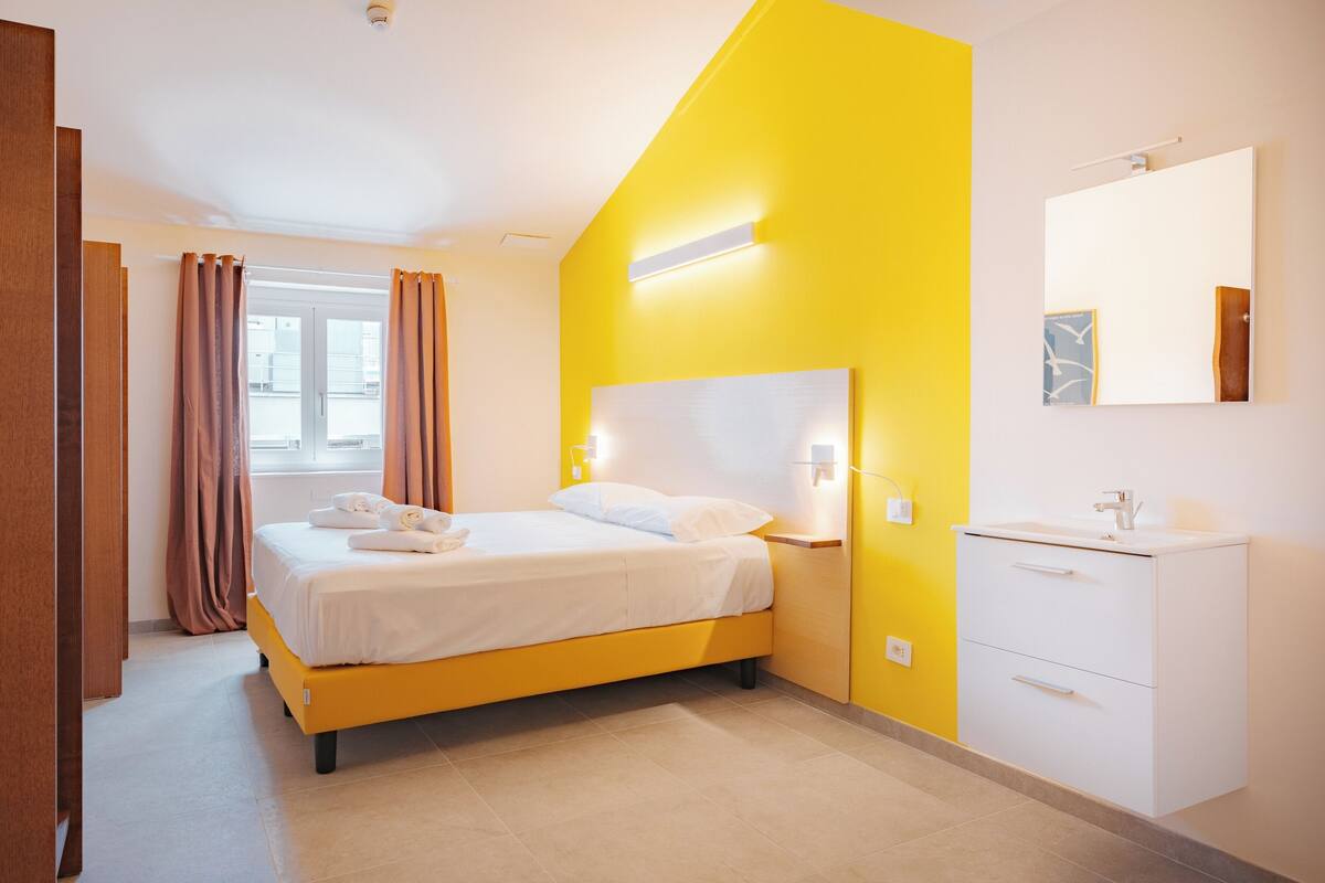 A modern hotel room with a bed, bedside tables, a mirror, and a window with curtains.