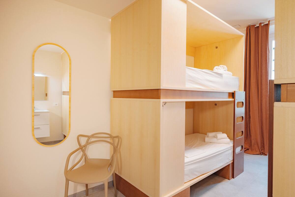 A bunk bed room with a wooden bunk bed, a chair, a mirror, and a bathroom visible in the background.