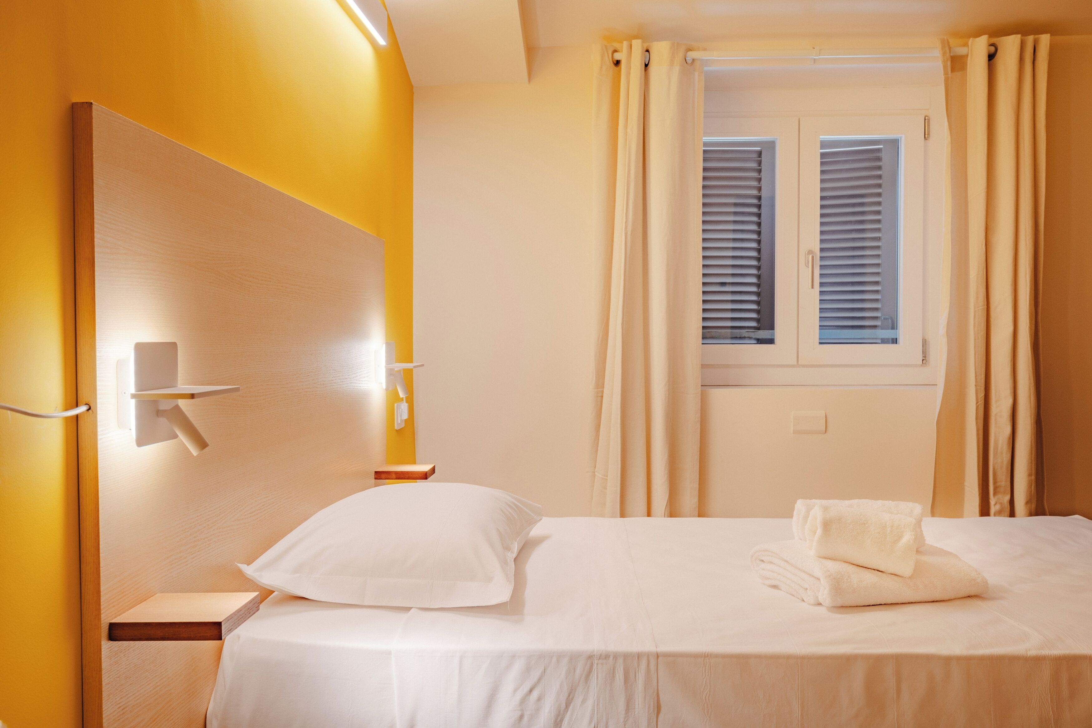 Habitación de hotel con cama, mesita de noche, luz de pared y ventana con cortinas.