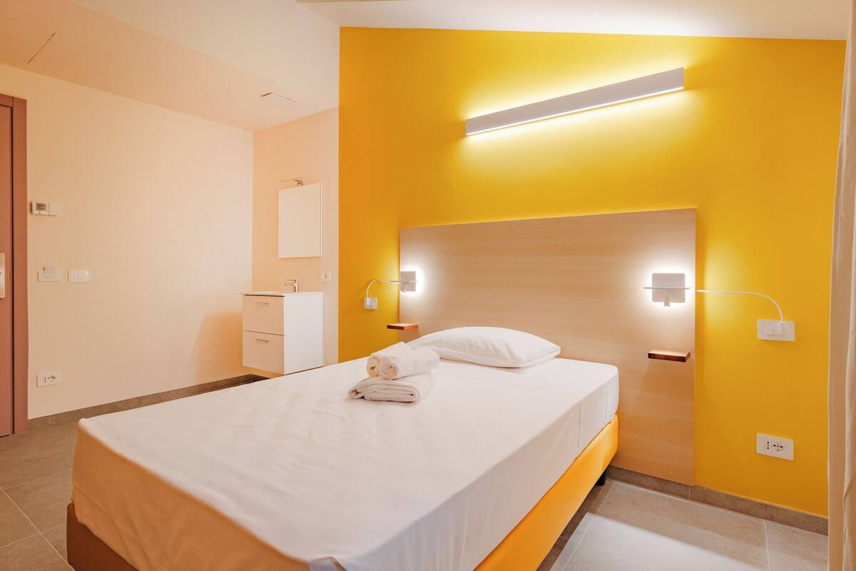 A modern hotel room with a bed, bedside tables, a nightstand, a mirror, and a wall-mounted light fixture.