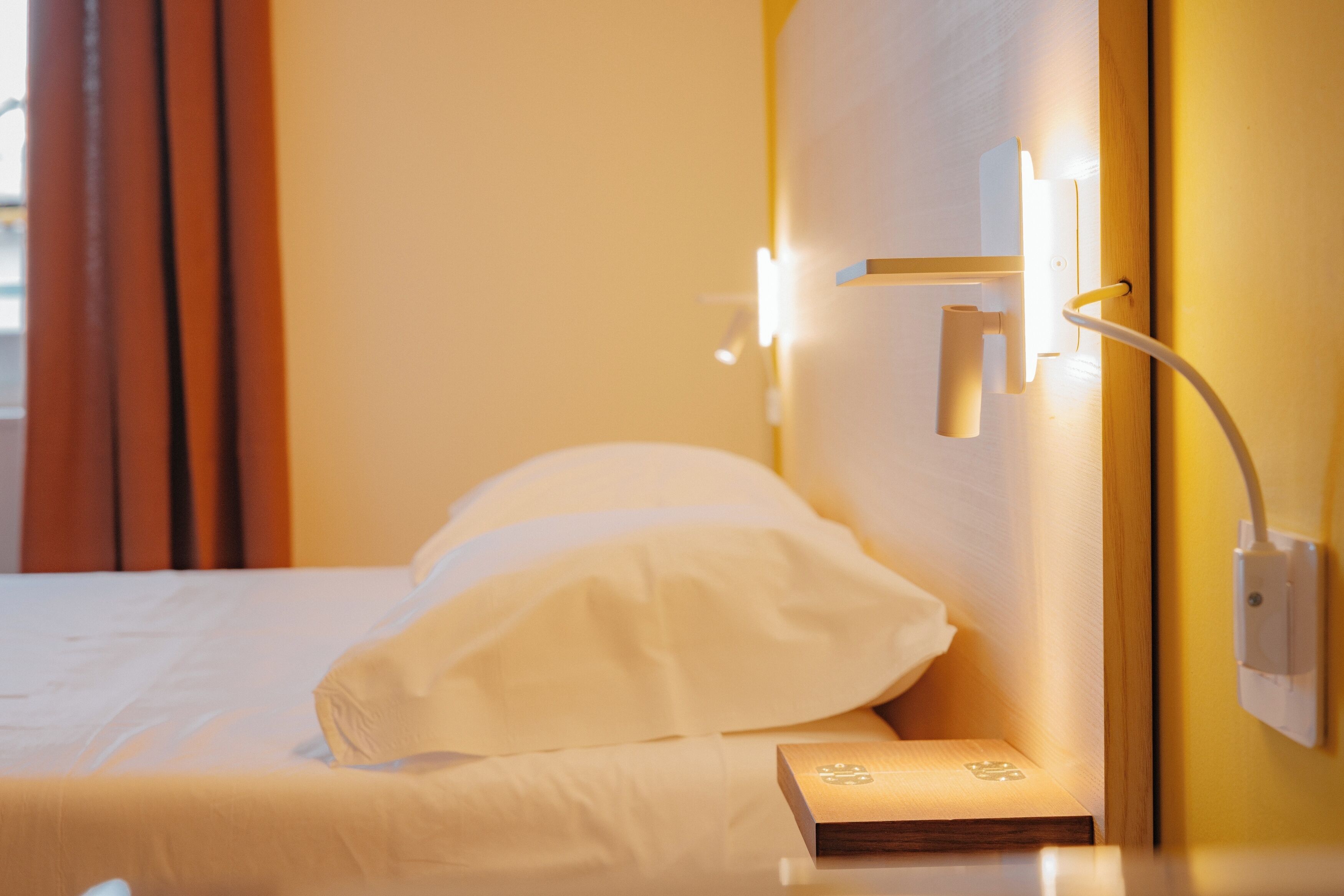 Una habitación de hotel con cama, mesita de noche, luz de pared y toma de corriente.