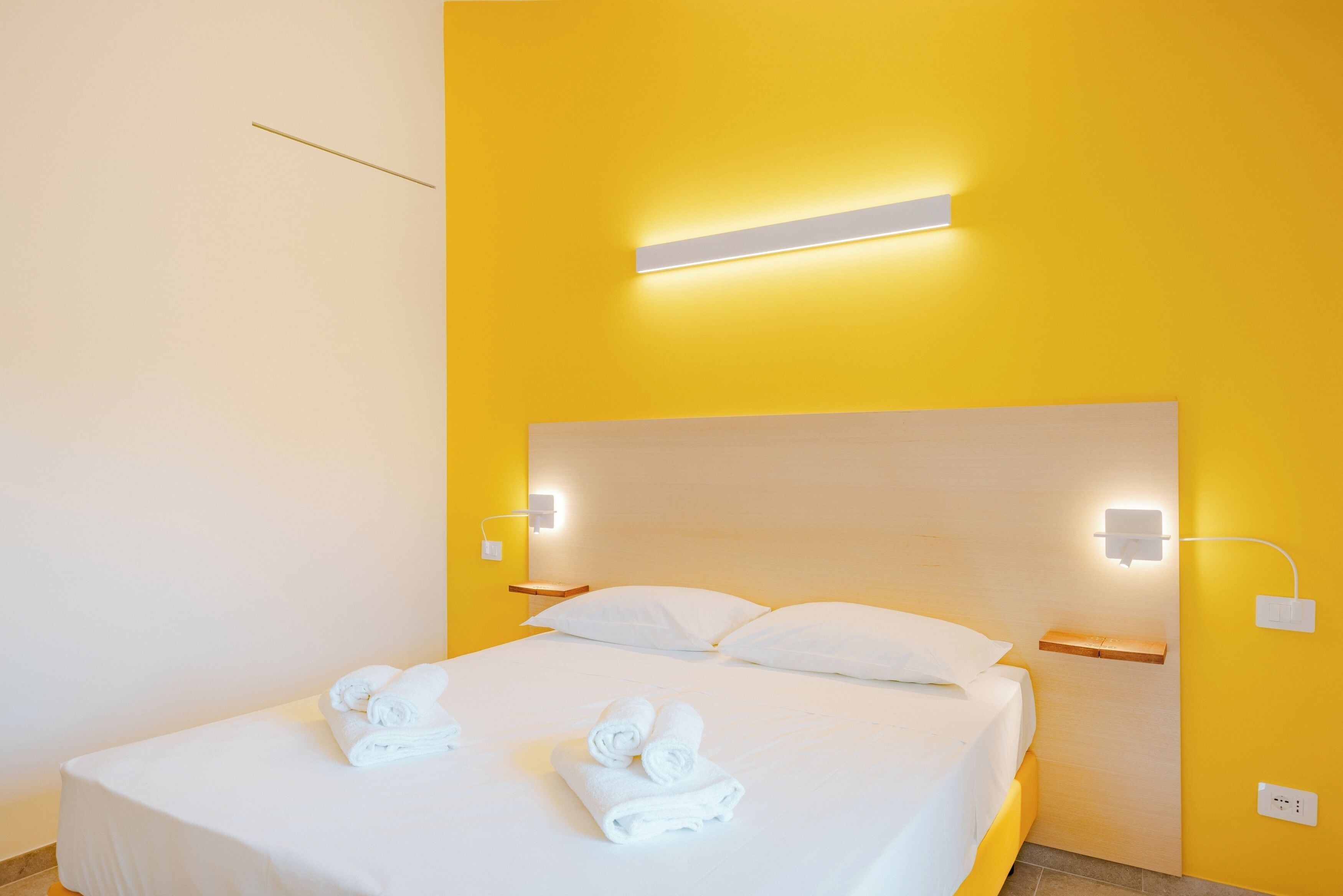 A hotel room with a bed, white linens, two wall-mounted light fixtures, and a bedside table.