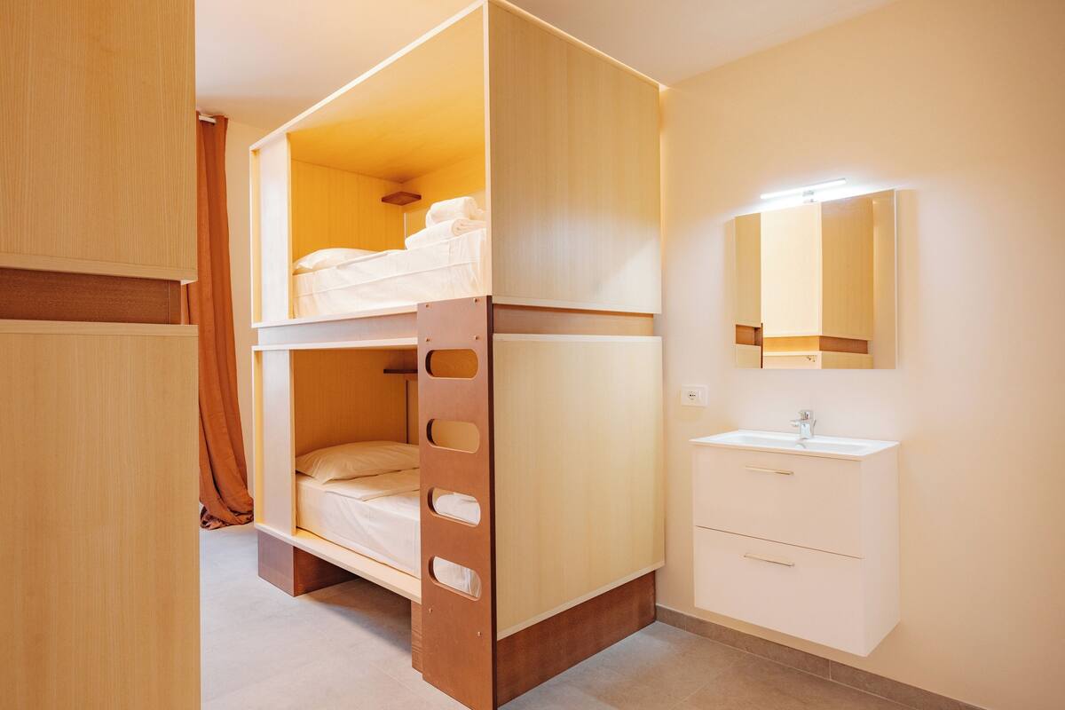 A bunk bed with a wooden frame, a white bedside table, a mirror above the sink, and a plain wall.