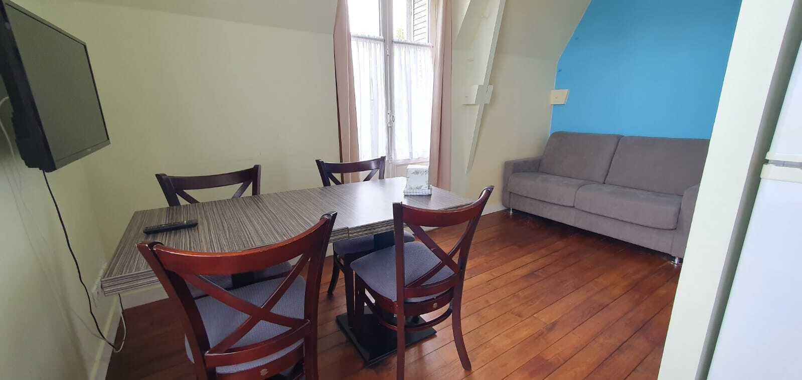 A dining area with a wooden table and four chairs, a grey sofa, and a TV mounted on the wall.