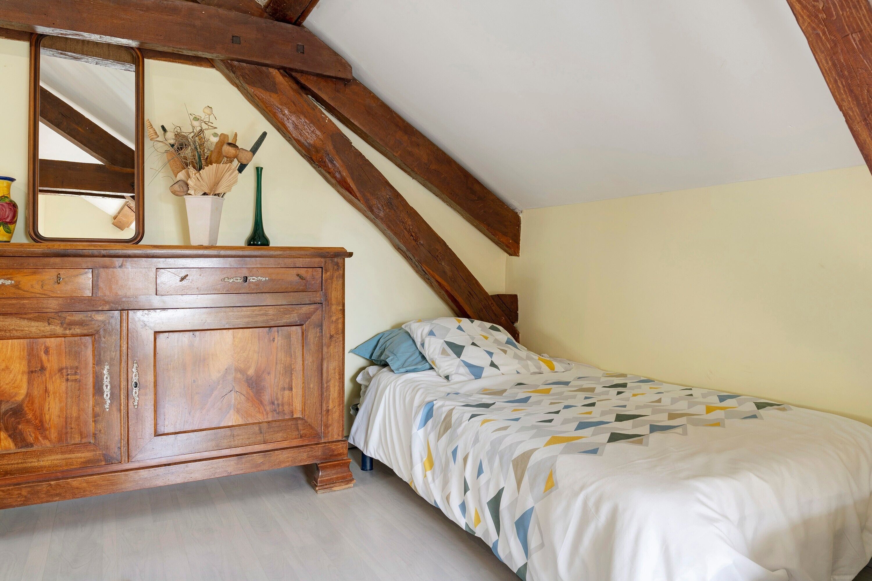 A bedroom with a wooden dresser, a bed with a geometric-patterned bedspread, a mirror, and a vase with dried flowers.