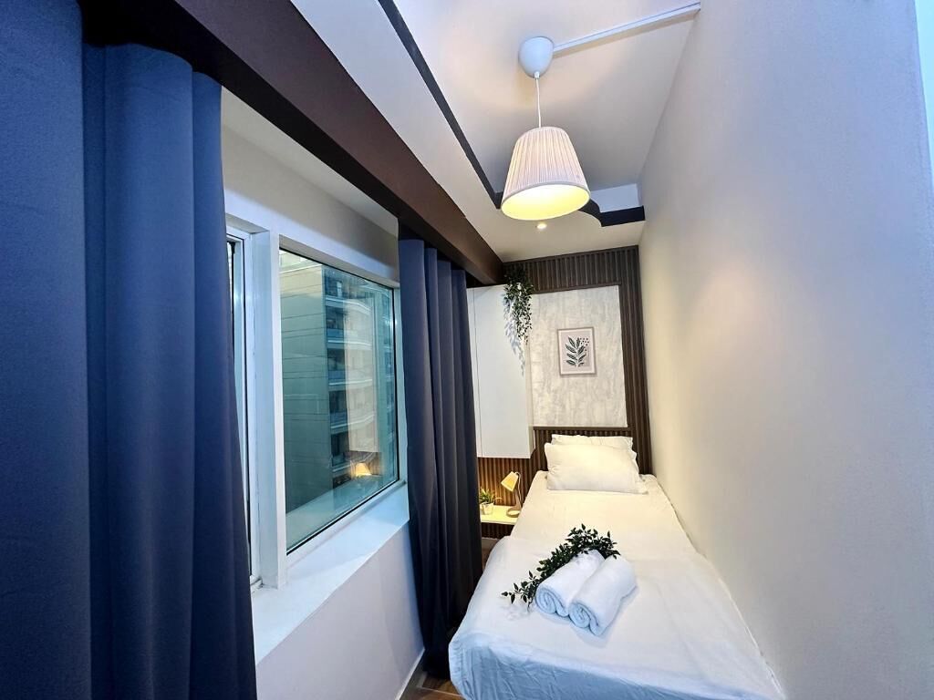 Una habitación de hotel moderna con un ventanal grande, una cama con ropa blanca, una mesita de noche con lámpara y un aplique de techo.