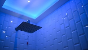 Eine moderne Dusche mit einem eckigen, perforierten Duschkopf, eingelassener Deckenbeleuchtung und weiß gefliesten Wänden mit geometrischem Muster.