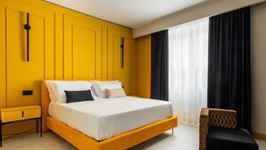 Una habitación de hotel moderna con una pared de acento amarilla, una cama grande, una mesita de noche, una silla y una ventana con cortinas translúcidas.