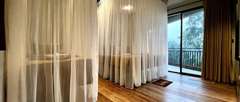 A room with a wooden floor, white curtains, and a balcony with a view of trees.