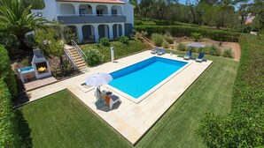 Ein Schwimmbad mit Liegestühlen und Grillplatz, umgeben von gepflegten Rasenflächen und einem großen Haus mit einem Ziegeldach.