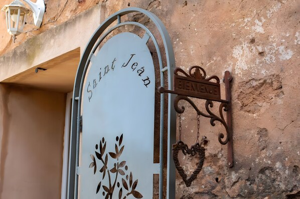 Une porte en métal sur laquelle est inscrit le mot « Saint Jean », une enseigne décorative en métal portant le nom « ELENZIE », accrochée au mur, et un petit ornement en forme de cœur.