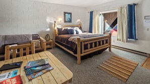 A cozy room with a wooden bed, a wooden table with magazines, a wooden chair, and a view of the outside through a window with curtains. - Château Rouge 107~ Cole Creek Corner (Red Lodge)