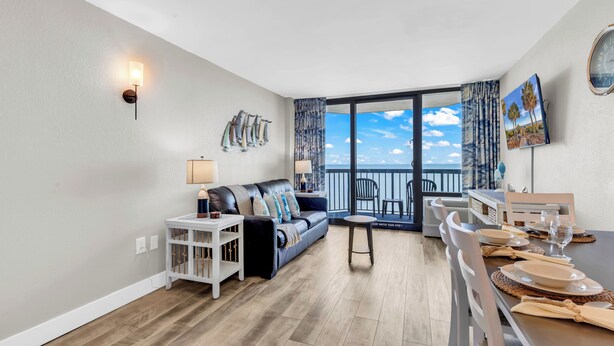 A modern living room with a sofa, dining table, and a balcony with a view. - Compass Cove Condos (Myrtle Beach)