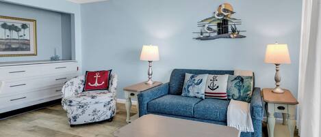 A living room with a sofa, two side tables with lamps, a coffee table, a chair with a red pillow, and a wall with a seascape painting.