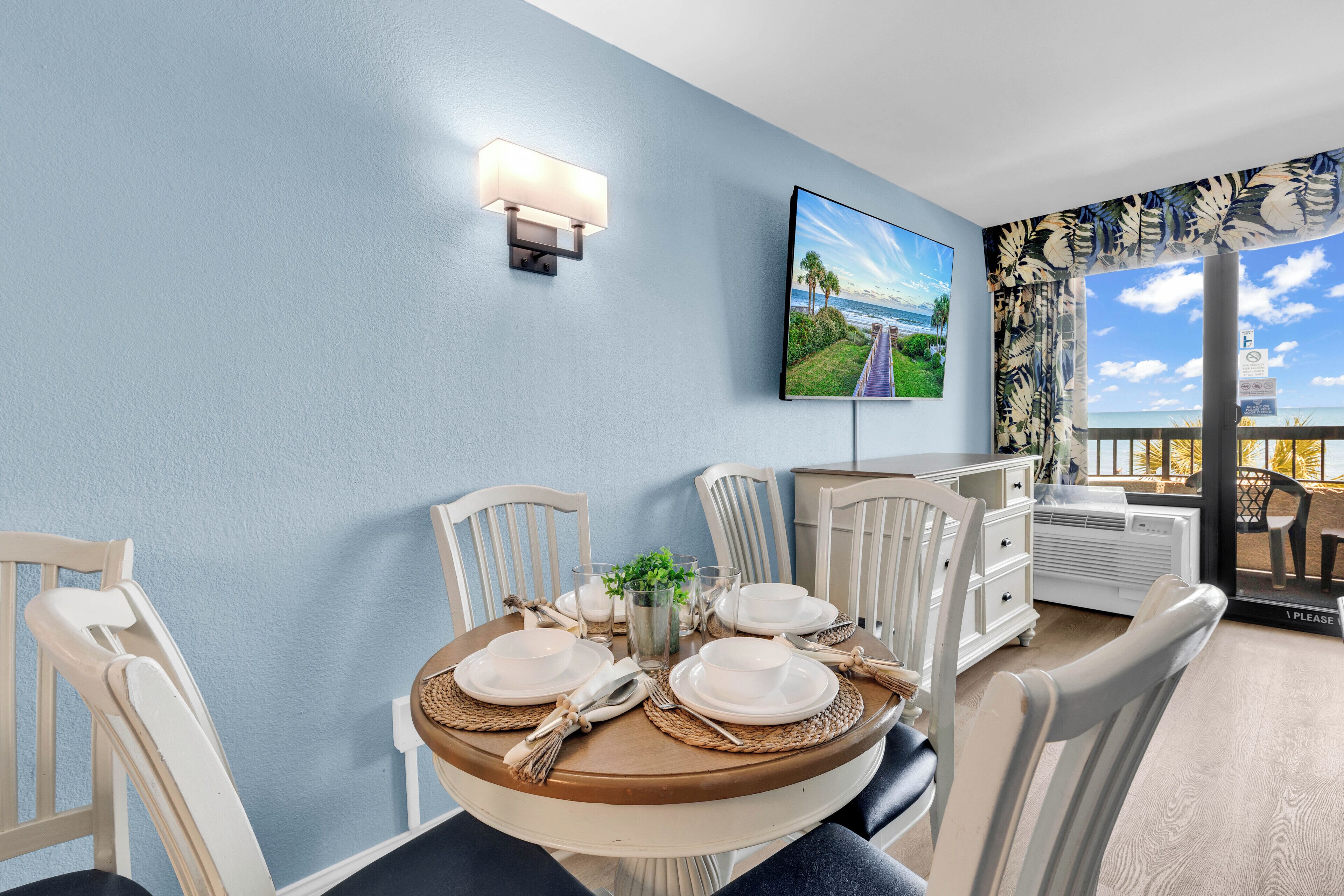 A dining area with a round table set for four, white chairs, a wall-mounted TV, and a view of the ocean through a window with a curtain.