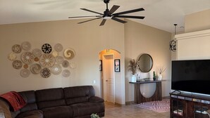 A living room with a brown leather sofa, a round wooden coffee table, a large flat-screen TV, and a ceiling fan.