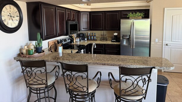 A kitchen with dark wood cabinets, a stainless steel refrigerator, a granite countertop, and a clock on the wall.