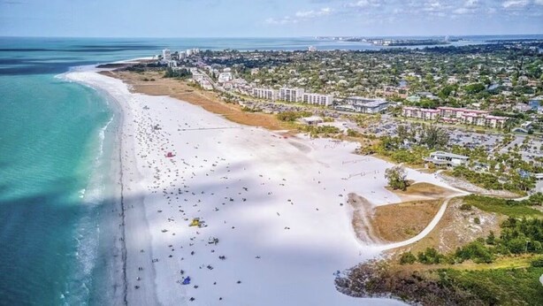 Vue aérienne d’une plage où l’on aperçoit de nombreux parasols, une étendue de sable et des bâtiments en arrière-plan.