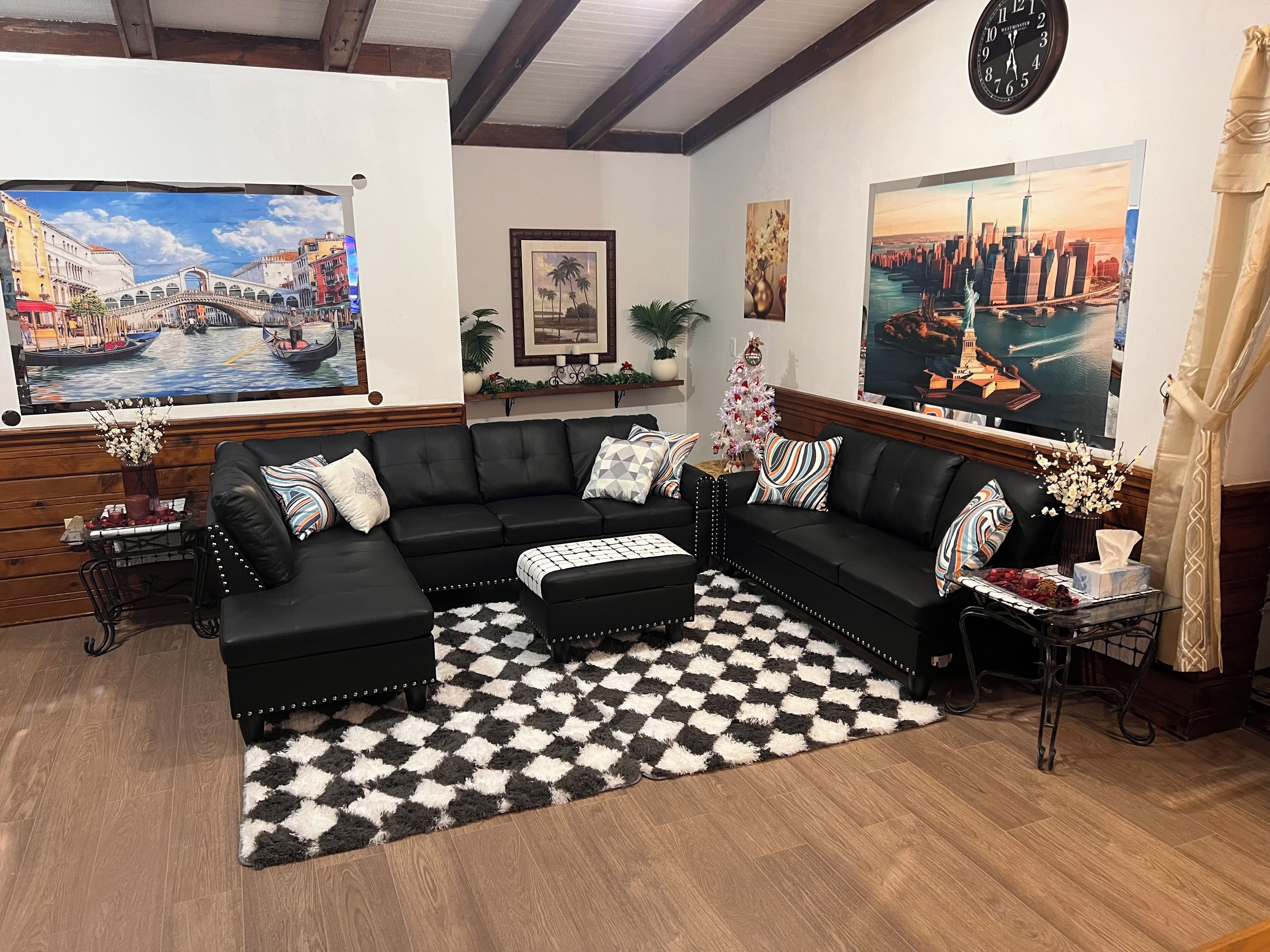A living room with a black leather sectional sofa, a checkered rug, and two paintings depicting cityscapes.
