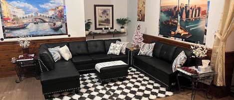 A living room with a black leather sectional sofa, a checkered rug, and two paintings depicting cityscapes.
