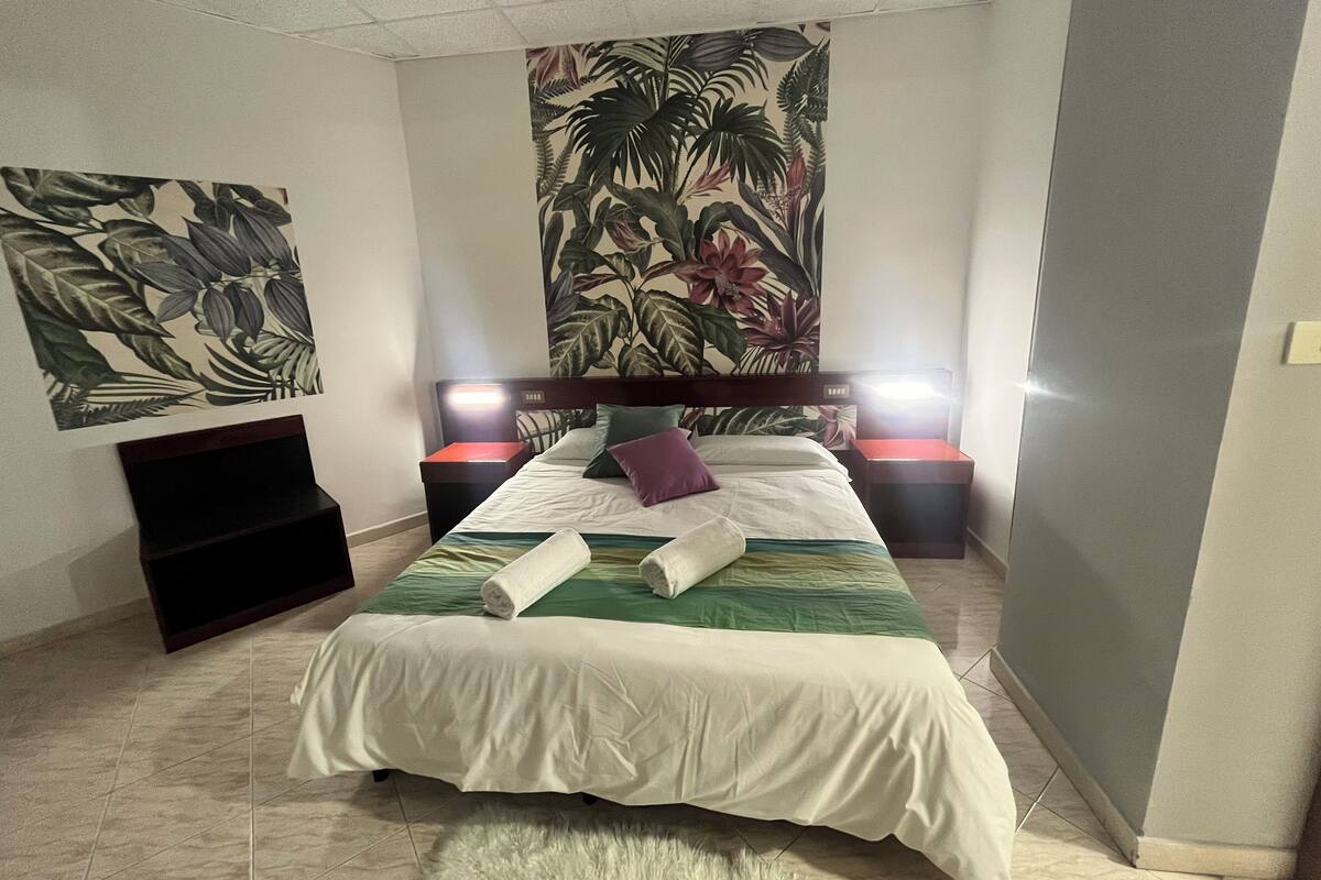 A bedroom with a bed, two bedside tables with lamps, a wall tapestry, and a small black shelf.