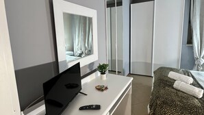 Ein modernes Schlafzimmer mit einem Bett, einem Schreibtisch mit Fernseher und Fernbedienung, einer kleinen Pflanze und einem Kleiderschrank.