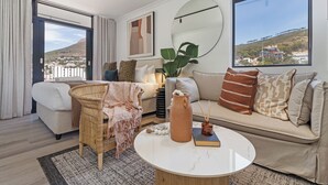 A modern living room with a sofa, a wicker chair, a coffee table, and a large window offering a view of a hillside. - The York  | Designer CBD Gem | Spacious Balcony (Cape Town)