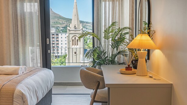 A hotel room with a bed, a desk with a lamp, a chair, and a view of a church and cityscape through large windows. - The York | Designer CBD Retreat (Cape Town)