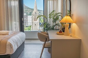 A hotel room with a bed, a desk with a lamp, a chair, and a view of a church and cityscape through large windows. - The York | Designer CBD Retreat (Cape Town)