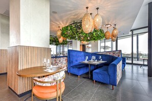 A modern restaurant with blue upholstered seating, a wooden table with orange cushions, and hanging wicker chandeliers. - Live Rosebank. Feel the Vibe. (Johannesburg)