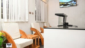 A reception area with a counter, two wooden chairs with cushions, a television mounted on the wall, and a plant on the left.