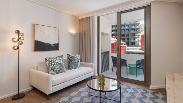 A modern living room with a sofa, a glass coffee table, and a floor lamp. There is a large window with a view of a building and a balcony. - Dolce Vita | Easy Coastal City Living (Cape Town)