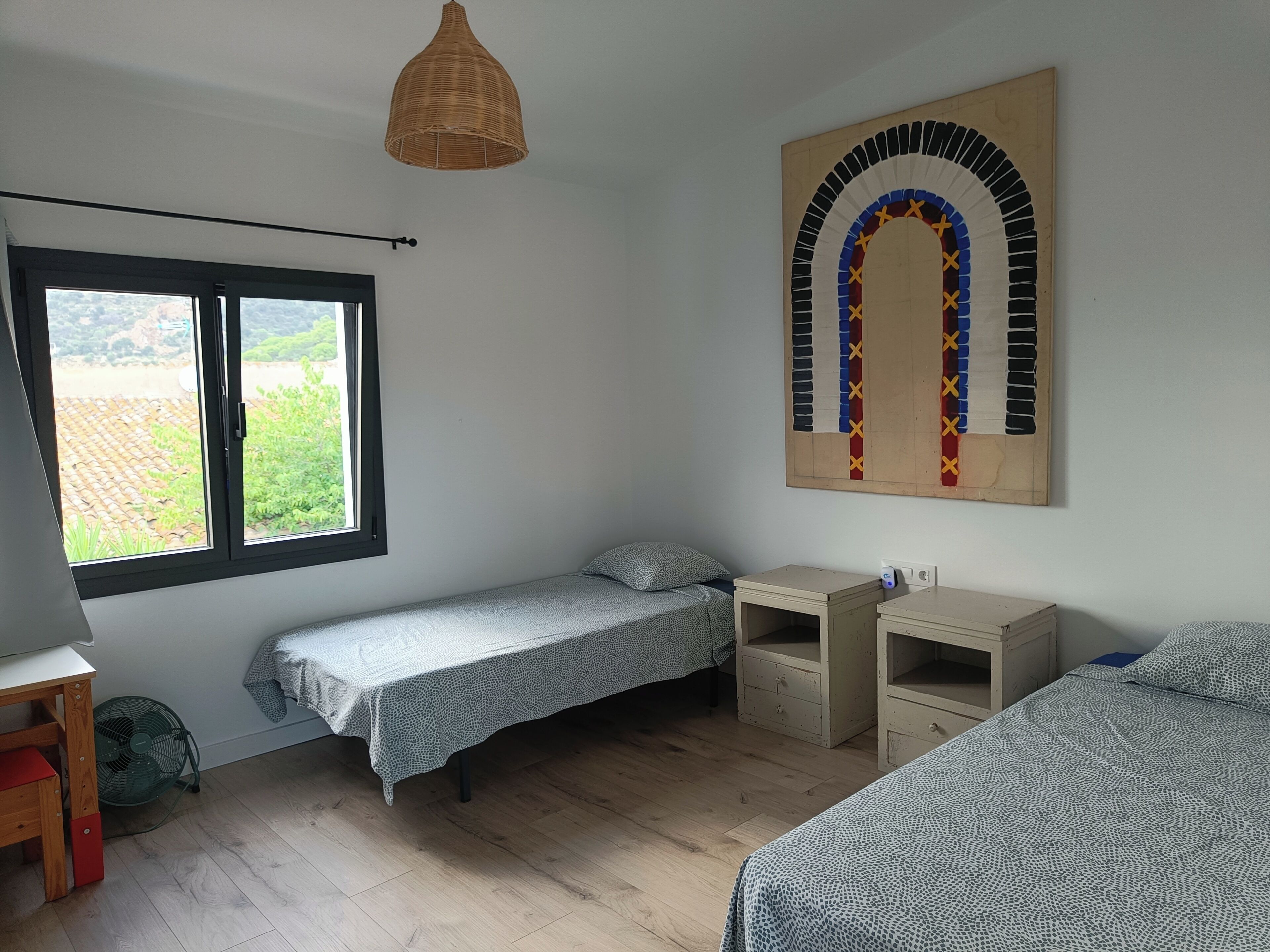 Een slaapkamer met twee eenpersoonsbedden, een bureau, een stoel, een staande ventilator en een groot raam.
