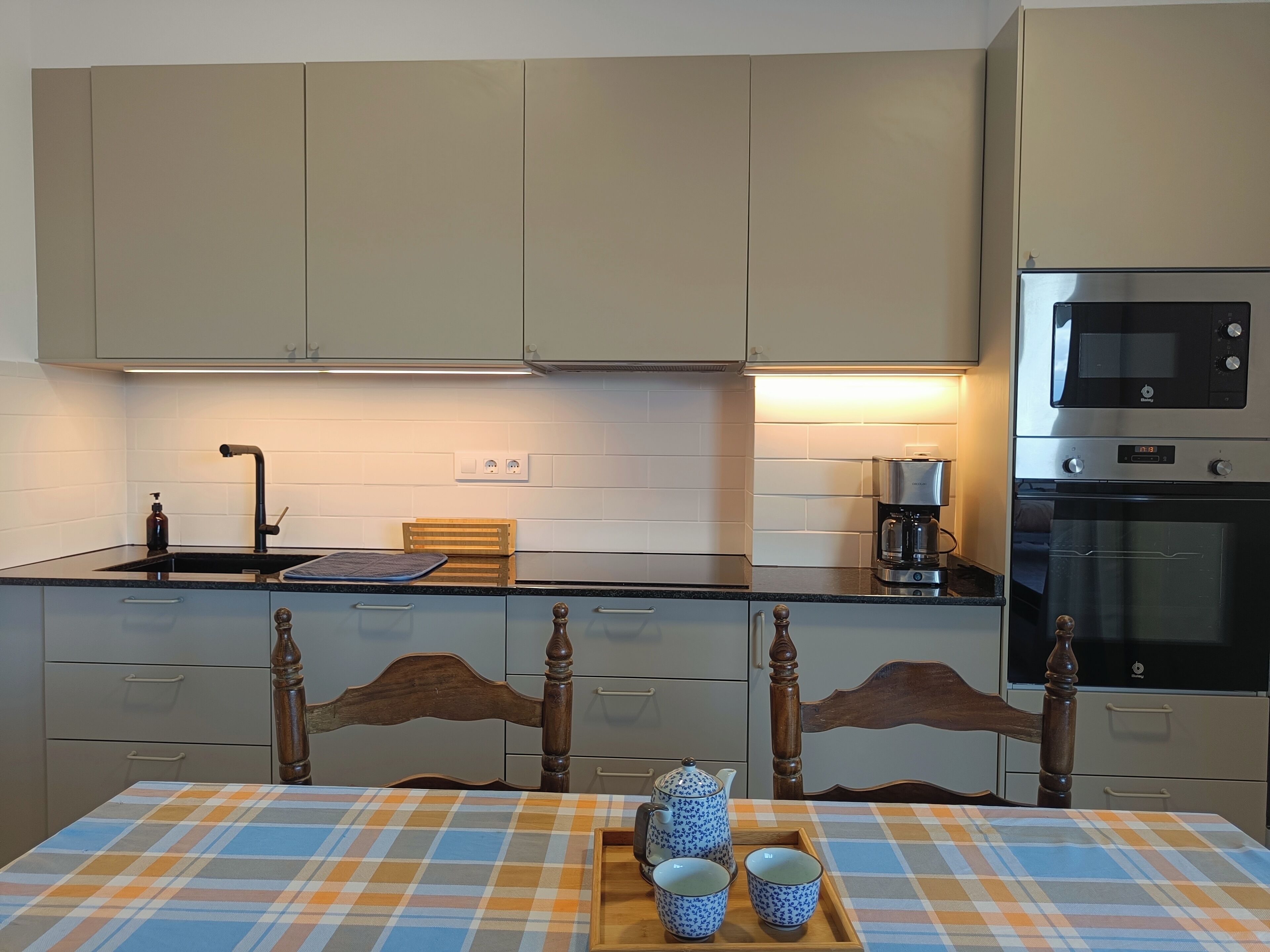 A modern kitchen with light-colored cabinets, a microwave, an oven, and a coffee maker. There is a wooden chair and a table with a plaid tablecloth.