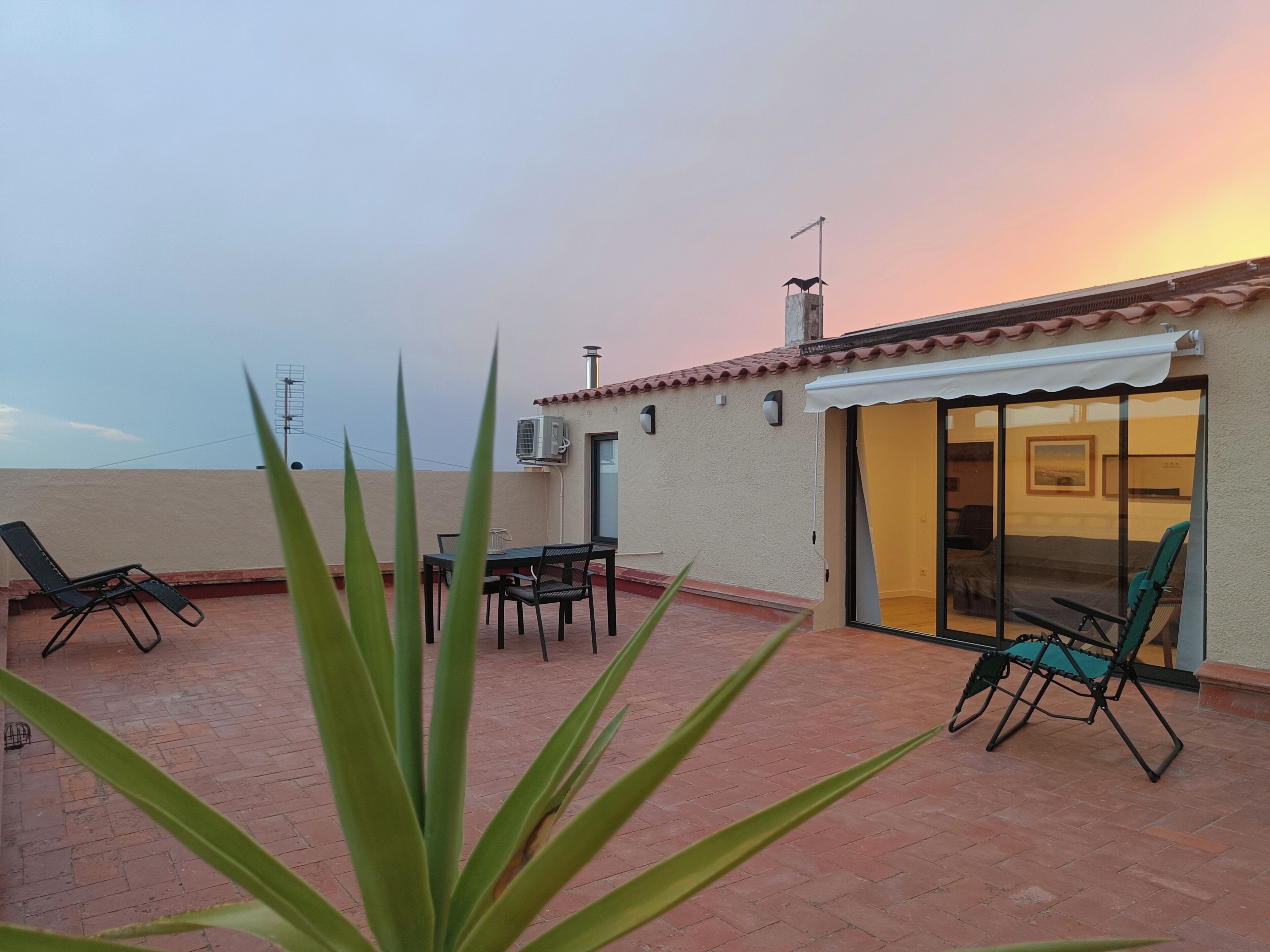 Eine Dachterrasse mit Tisch und Stühlen, einer Schiebetür und Blick auf den Sonnenuntergang.