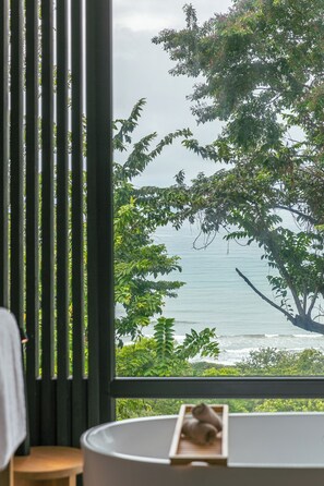 Ein Zimmer mit großem Fenster mit Blick auf Strand und Meer. In der Nähe des Fensters befinden sich ein Holzschemel und ein weißer Stuhl.