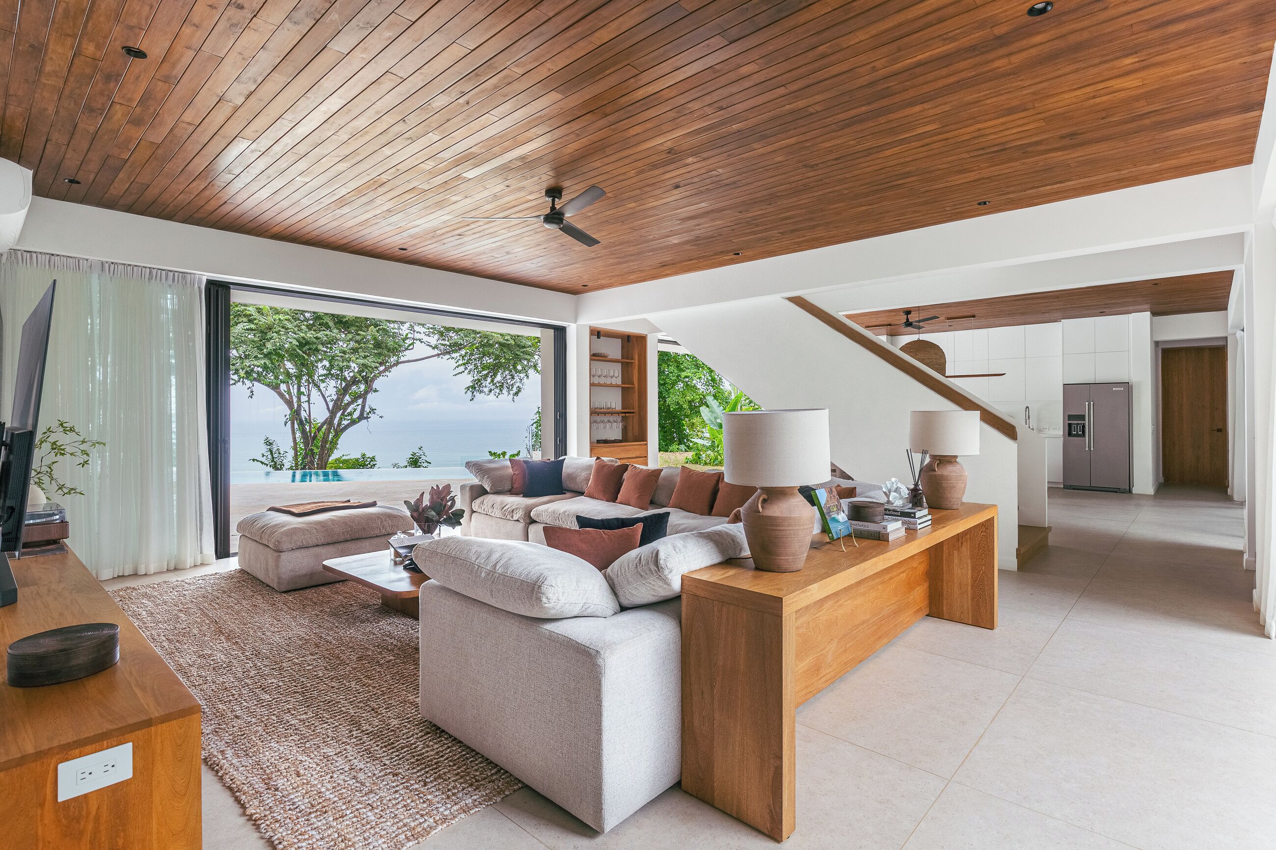 A modern living room with a wooden ceiling, a large window with a view of the sea, a sofa set, and a coffee table.