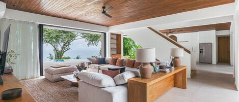 A modern living room with a wooden ceiling, a large window with a view of the sea, a sofa set, and a coffee table.