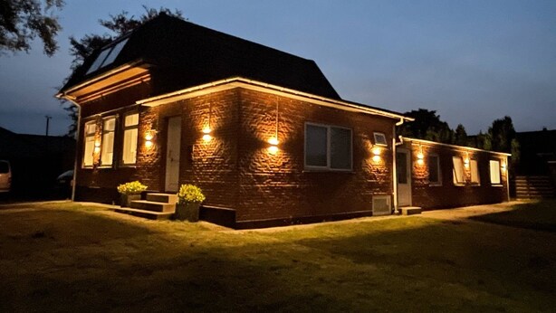 A brick house with a sloped roof, illuminated by exterior lights, and a small front porch with steps leading to the door.
