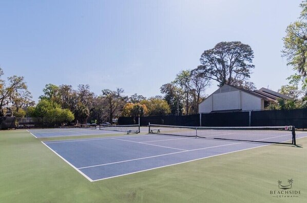 Una cancha de tenis con una red en el centro, rodeada de árboles y un edificio al fondo.