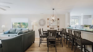 A modern living room with a grey sofa, wooden dining table, and chairs. There is a kitchen area with a white countertop and cabinets. - 70 Springwood Villas - 3BR/3 Bath (Hilton Head Island, SC)