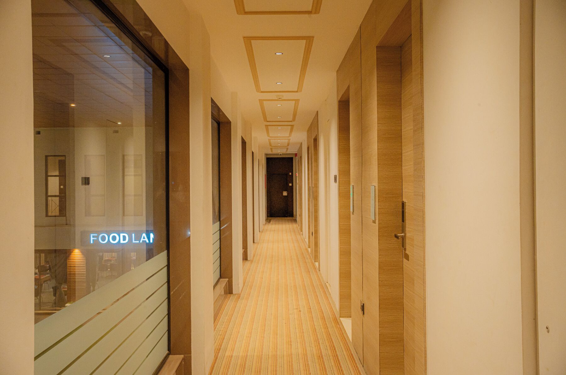 A hotel hallway with a wooden floor, a glass wall on the left, and a door on the right.