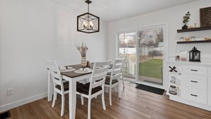 A dining area with a hexagonal ceiling light, white chairs, a wooden table, and a view through a glass door to a backyard. - 3 bed,2 bath Luxury & Private Turf Yard (Rapid City)