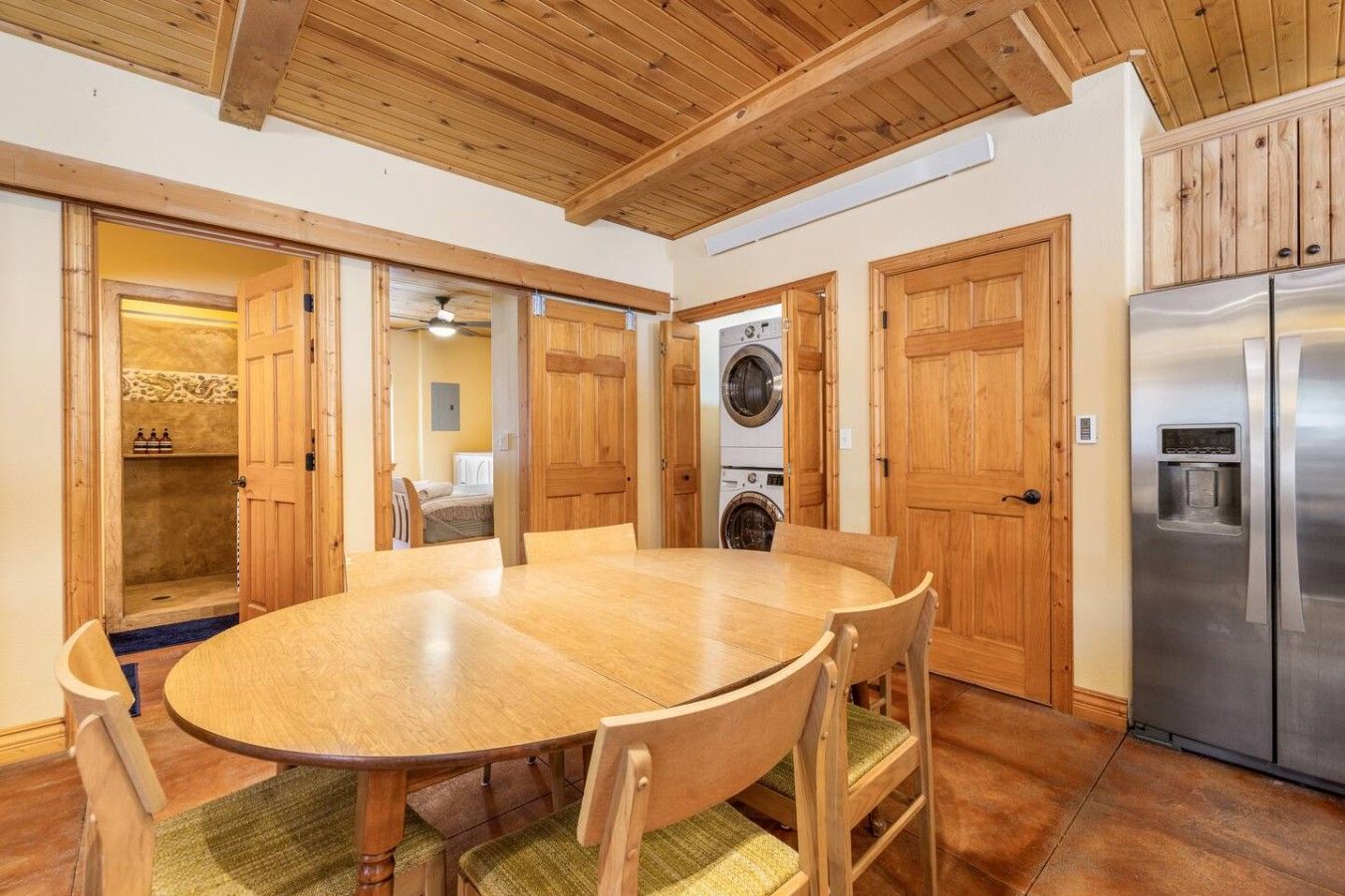 Une table de salle à manger en bois, avec des chaises, un lave-linge et un réfrigérateur, dans une pièce dont le plafond et les portes sont en bois.