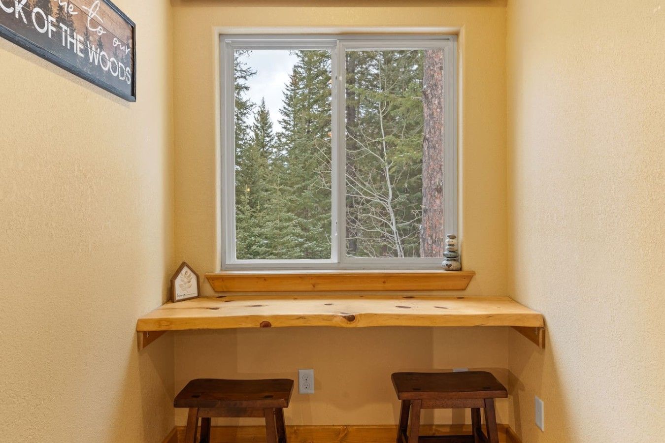 Quarto de canto com janela para uma floresta, um banco de madeira e uma mesa pequena.